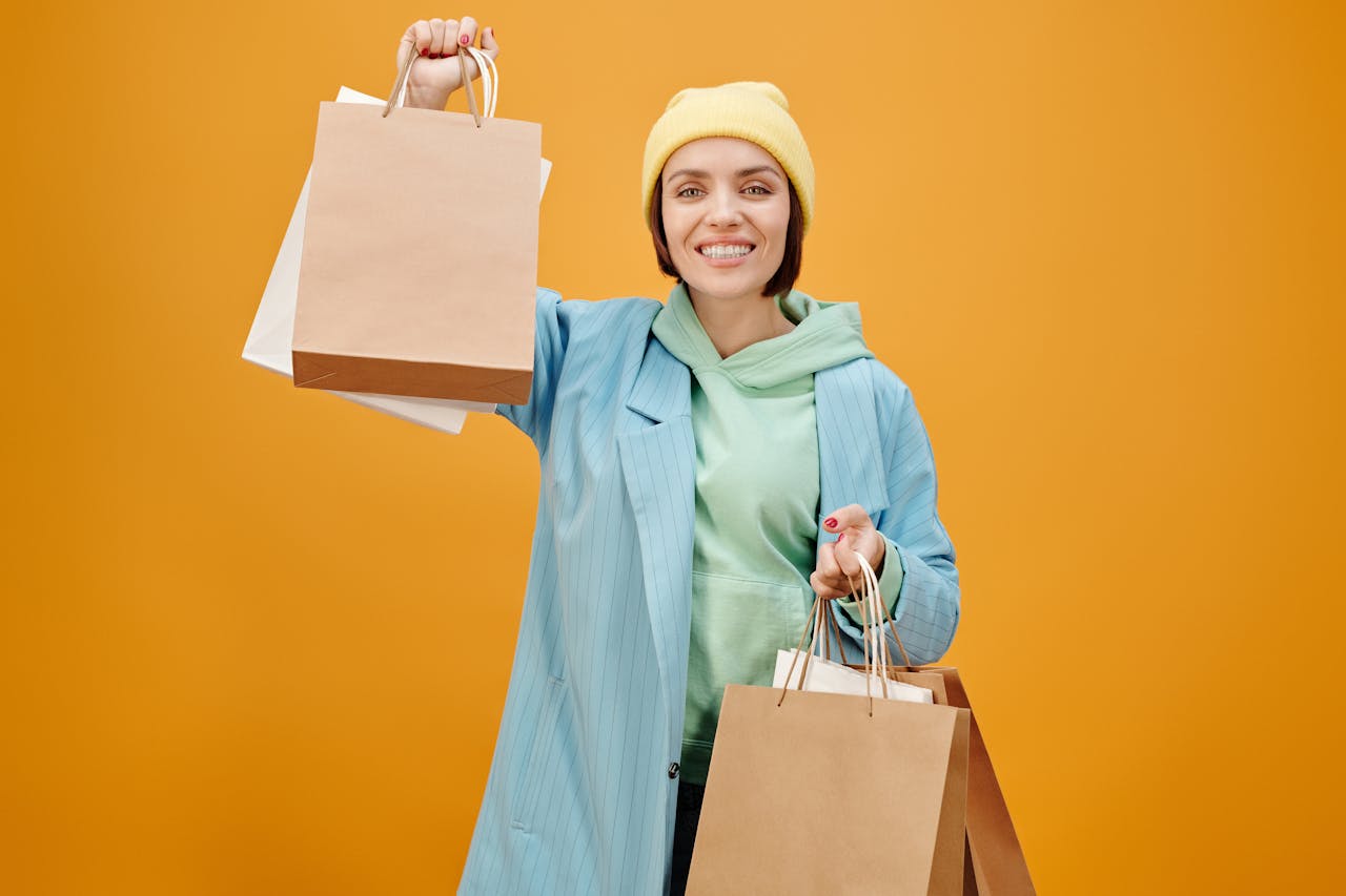 A cheerful woman in a beanie and blue coat holding shopping bags, against a vibrant yellow background.